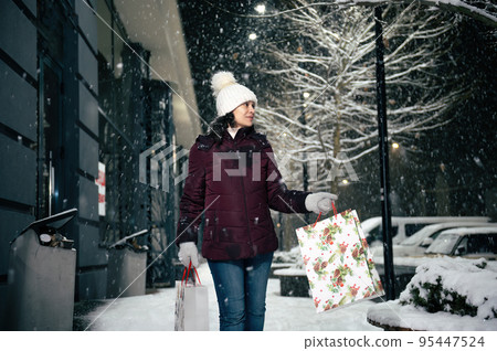 Delightful young multi-ethnic woman walking down the street with shopping bags, on a snowy winter cool evening. Merry Christmas. 95447524