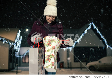 Multi ethnic pretty woman wearing dawn jacket, wool mittens and hat, with shopping bags walking down the snow covered street on a snowy winter cold evening. Merry Christmas and Happy New Year concept 95447525