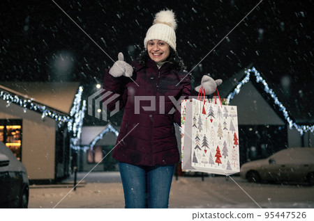 Charming young woman in white wool hat and mittens, experiencing happiness, showing a thumbs up, smiling cheerfully to the camera, standing against a snow covered pine tree on a snowy winter evening Charming young woman in white wool hat and mittens, experiencing happiness, showing a thumbs up, smiling cheerfully to the camera, standing against a snow covered pine tree on a snowy winter evening 95447526