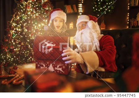 Santa Claus surprising little girl while open magical box near Christmas tree. New Year concept Santa Claus surprising little girl while open magical box near Christmas tree. New Year concept 95447745