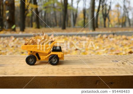 A small toy yellow truck is loaded with yellow fallen leaves. The car stands on a wooden surface against a background of a blurry autumn park. Cleaning and removal of fallen leaves. Seasonal works A small toy yellow truck is loaded with yellow fallen leaves. The car stands on a wooden surface against a background of a blurry autumn park. Cleaning and removal of fallen leaves. Seasonal works 95448483
