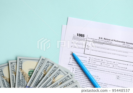 1065 tax form lies near hundred dollar bills and blue pen on a light blue background. US Return for parentship income 1065 tax form lies near hundred dollar bills and blue pen on a light blue background. US Return for parentship income 95448537