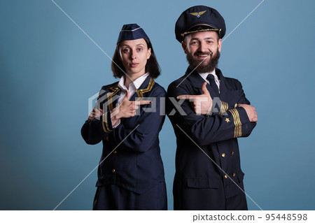 Portrait of airplane capitan and flight attendant pointing at each other while standing in studio with blue background. Aviation crew wearing professional uniform, medium shot Portrait of airplane capitan and flight attendant pointing at each other while standing in studio with blue background. Aviation crew wearing professional uniform, medium shot 95448598