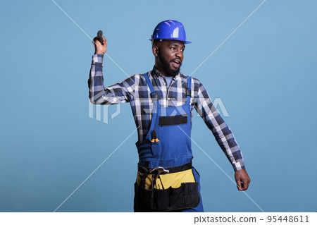 African american construction worker wearing coverall and tool belt annoyingly throwing hammer. Stressed aggressive construction worker tired of so much work throwing tools in anger. 95448611