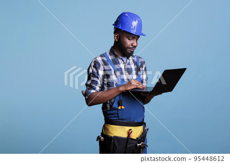 Construction worker in work uniform and hard hat using modern laptop computer. Professional male construction site supervisor wearing a tool belt while using a computer on a blue background. 95448612