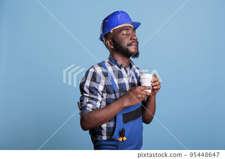 Relaxed construction worker enjoying a cup of favorite coffee before starting work on an interior renovation. Builder wearing coveralls and hard hat against blue background in studio shot. Relaxed construction worker enjoying a cup of favorite coffee before starting work on an interior renovation. Builder wearing coveralls and hard hat against blue background in studio shot. 95448647