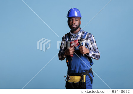 Professional construction worker with coveralls and work tools against blue background. Studio shot african american man looking at camera holding electric cordless drill. Professional construction worker with coveralls and work tools against blue background. Studio shot african american man looking at camera holding electric cordless drill. 95448652