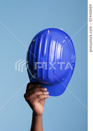 Construction worker holding a protective helmet in a studio shot on blue background. Unrecognizable person showing on camera object of occupational protection, concept of safety in construction. 95448664