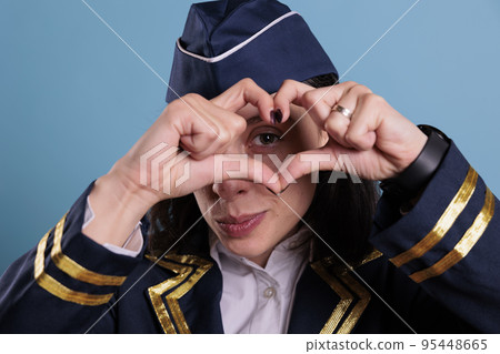 Smiling airliner stewardess showing heart shaped love symbol with fingers, conceptual romance gesture. Friendly air hostess in aviation uniform expressing affection feelings front view medium shot 95448665