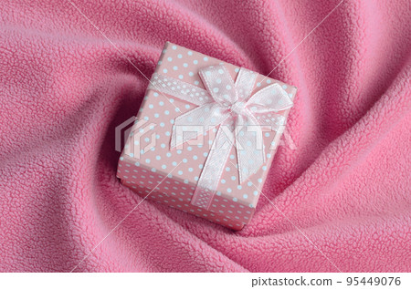 A small gift box in pink with a small bow lies on a blanket of soft and furry light pink fleece fabric with a lot of relief folds. Packing for a gift to your lovely girlfriend A small gift box in pink with a small bow lies on a blanket of soft and furry light pink fleece fabric with a lot of relief folds. Packing for a gift to your lovely girlfriend 95449076