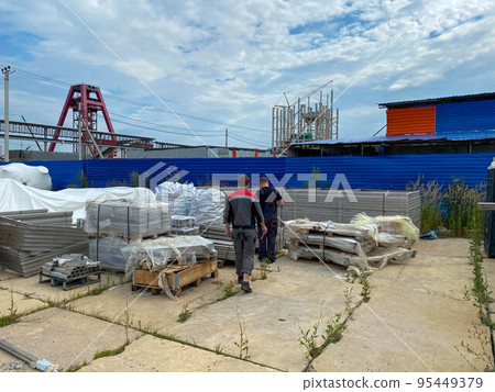 Male workers walk through a warehouse of industrial equipment and materials in boxes in an open-air storage area. View from the back Male workers walk through a warehouse of industrial equipment and materials in boxes in an open-air storage area. View from the back 95449379