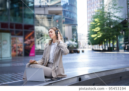 Smiling businesswoman relaxing, listetning to music in headphones from her laptop, sitting alone on street of city centre, wearing suit Smiling businesswoman relaxing, listetning to music in headphones from her laptop, sitting alone on street of city centre, wearing suit 95453194