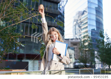 Enthusiastic businesswoman feels upbeat after morning cup of coffee, raise her cup up and dances outside Enthusiastic businesswoman feels upbeat after morning cup of coffee, raise her cup up and dances outside 95453195