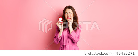 Beautiful girl dream of her wedding day, looking thoughtful up, showing engagement ring in red box and smiling, standing over pink background Beautiful girl dream of her wedding day, looking thoughtful up, showing engagement ring in red box and smiling, standing over pink background 95453271