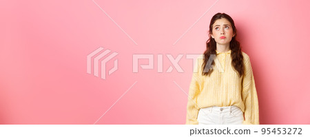 Image of upset and gloomy young woman pulling sad face, looking up with regret or jealousy, standing disappointed against pink background Image of upset and gloomy young woman pulling sad face, looking up with regret or jealousy, standing disappointed against pink background 95453272