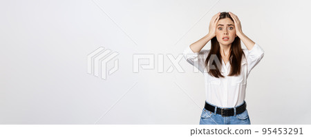Worried and shocked young woman, looking stunned at disaster, holding hands on head, panicking, standing over white background 95453291