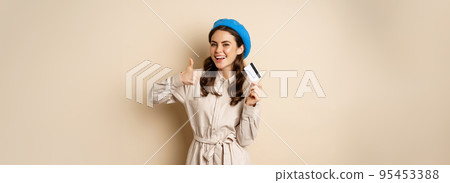 Concept of shopping and sale. Happy young woman in stylish clothes, showing credit card store discount and thumbs up, recommending, standing over beige background Concept of shopping and sale. Happy young woman in stylish clothes, showing credit card store discount and thumbs up, recommending, standing over beige background 95453388