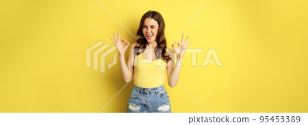 Smiling attractive woman showing okay signs and winking, assure you, praise and compliment smth good, recommending, standing over yellow background Smiling attractive woman showing okay signs and winking, assure you, praise and compliment smth good, recommending, standing over yellow background 95453389