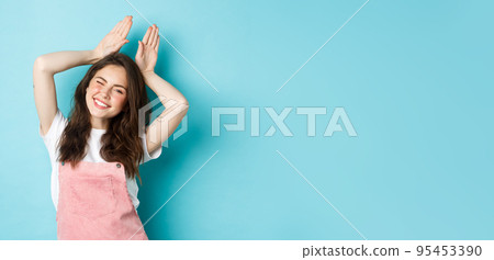 Cheerful girl celebrating Easter, showing easter bunny gesture and smiling cute at camera, standing over blue background in spring outfit 95453390