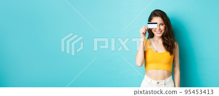 Young stylish woman showing plastic credit card over eye, looking pleased adn confident, smiling at camera, standing over blue background 95453413