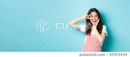 Portrait of excited attractive woman showing v-sign near eye, making kawaii gesture and smiling happy at camera, standing upbeat against blue background Portrait of excited attractive woman showing v-sign near eye, making kawaii gesture and smiling happy at camera, standing upbeat against blue background 95453434