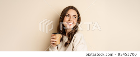 Takeaway and cafe concept. Beautiful feminine woman smiling, holding cup of coffee, posing against beige background 95453700
