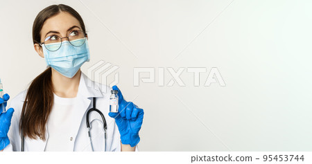 Covid-19 vaccination and healthcare concept. Young doctor in medical mask showing syringe and vaccine from coronavirus omicron variant, standing over white background 95453744