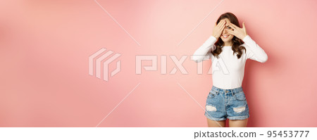 Excited glamour girlfriend peeking through fingers with surprised, yearning face, seeing gifts or presents, standing over pink background 95453777