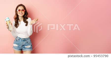 Stylish girl in spring outfit, wearing sunglasses, holding water bottle with lemon, healthy drink, laughing and smiling, standing over pink background Stylish girl in spring outfit, wearing sunglasses, holding water bottle with lemon, healthy drink, laughing and smiling, standing over pink background 95453790
