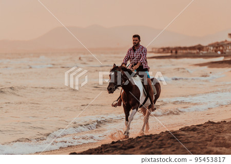 A modern man in summer clothes enjoys riding a horse on a beautiful sandy beach at sunset. Selective focus A modern man in summer clothes enjoys riding a horse on a beautiful sandy beach at sunset. Selective focus 95453817