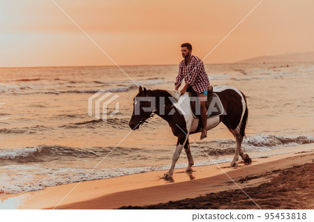 A modern man in summer clothes enjoys riding a horse on a beautiful sandy beach at sunset. Selective focus A modern man in summer clothes enjoys riding a horse on a beautiful sandy beach at sunset. Selective focus 95453818