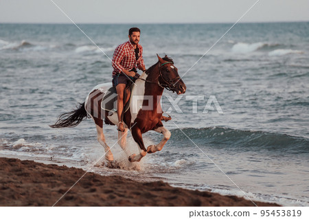 A modern man in summer clothes enjoys riding a horse on a beautiful sandy beach at sunset. Selective focus A modern man in summer clothes enjoys riding a horse on a beautiful sandy beach at sunset. Selective focus 95453819