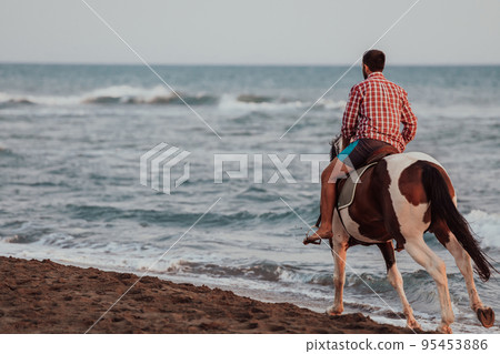 A modern man in summer clothes enjoys riding a horse on a beautiful sandy beach at sunset. Selective focus  95453886