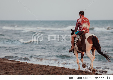 A modern man in summer clothes enjoys riding a horse on a beautiful sandy beach at sunset. Selective focus  95453914