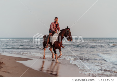 A modern man in summer clothes enjoys riding a horse on a beautiful sandy beach at sunset. Selective focus A modern man in summer clothes enjoys riding a horse on a beautiful sandy beach at sunset. Selective focus 95453978