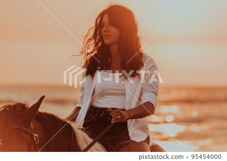 Woman in summer clothes enjoys riding a horse on a beautiful sandy beach at sunset. Selective focus Woman in summer clothes enjoys riding a horse on a beautiful sandy beach at sunset. Selective focus 95454000
