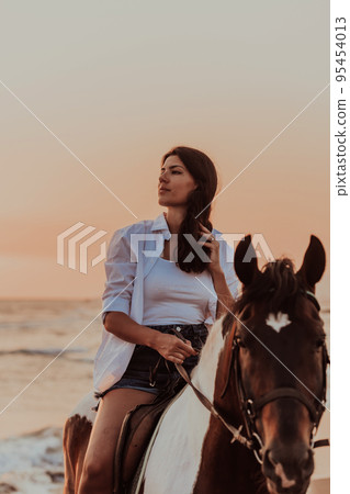 Woman in summer clothes enjoys riding a horse on a beautiful sandy beach at sunset. Selective focus Woman in summer clothes enjoys riding a horse on a beautiful sandy beach at sunset. Selective focus 95454013