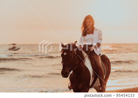 Woman in summer clothes enjoys riding a horse on a beautiful sandy beach at sunset. Selective focus Woman in summer clothes enjoys riding a horse on a beautiful sandy beach at sunset. Selective focus 95454133