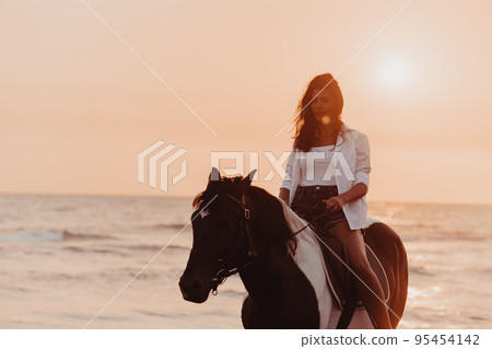 Woman in summer clothes enjoys riding a horse on a beautiful sandy beach at sunset. Selective focus  95454142