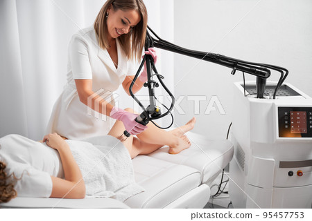 Dermatologist working with modern laser device for polishing and repairing skin defects. Beautician demonstrating work of CO2 laser, warming skin of clients thigh, stimulating local rejuvenation. 95457753