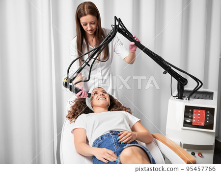 Visiting beautician to treat post-traumatic acne scars. Female dermatologist performing laser resurfacing on face skin of smiling young lady. 95457766