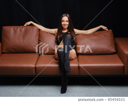 Positive sportive woman sits on leather sofa in warm socks spreading hands into the sides 95459331