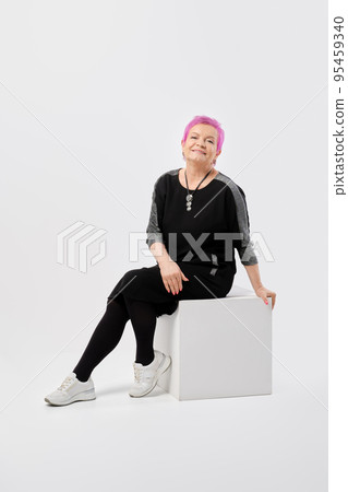 Happy old woman with short pink hair in long knitted dress sits on box Happy old woman with short pink hair in long knitted dress sits on box 95459340