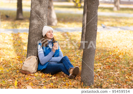 Autumn chill out. Caucasian woman in warm casual wear sits under a tree with coffee in hand in the park outdoor in golden fall, selected focus. 95459554