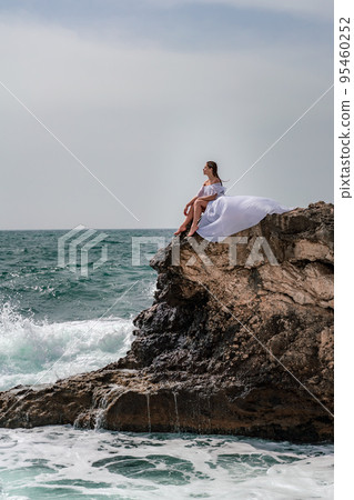 A woman in a storm sits on a stone in the sea. Dressed in a white long dress, waves crash against the rocks and white spray rises. A woman in a storm sits on a stone in the sea. Dressed in a white long dress, waves crash against the rocks and white spray rises. 95460252
