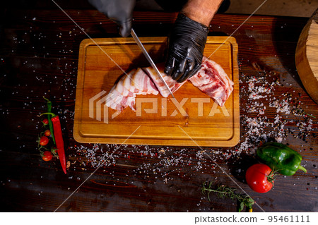 Salted Lard, Raw Pork with Spices on Wooden Cutting Board Studio Photo. A butcher in black gloves thinly slices lard 95461111