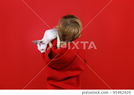 a boy in a red suit stands on a red background, turned his back, holds a small white kitten on his shoulder, which looks around 95462920