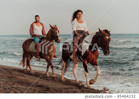 A loving couple in summer clothes riding a horse on a sandy beach at sunset. Sea and sunset in the background. Selective focus A loving couple in summer clothes riding a horse on a sandy beach at sunset. Sea and sunset in the background. Selective focus 95464132