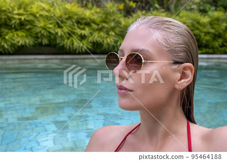 Side view portrait of confident sexy woman in pool looking away. Woman with wet hair wearing sunglasses and red swimsuit against plants on sunny summer day. 95464188