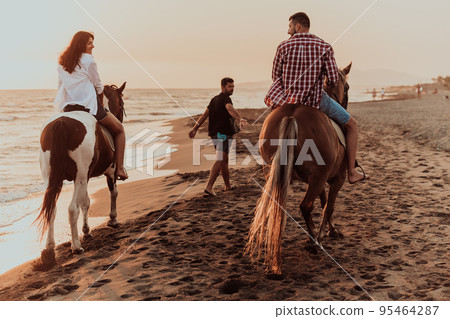 A loving couple in summer clothes riding a horse on a sandy beach at sunset. Sea and sunset in the background. Selective focus A loving couple in summer clothes riding a horse on a sandy beach at sunset. Sea and sunset in the background. Selective focus 95464287